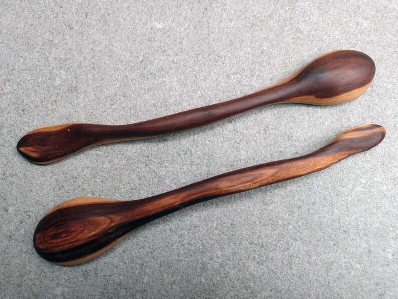 May include: Two handcrafted wooden spoons featuring a distinct two-tone color palette with deep dark brown and lighter tan wood grain patterns. Each spoon has a long, curved handle and a smooth, rounded bowl, showcasing natural wood textures.