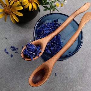May include: Two light brown wooden spoons with smooth, curved handles. One spoon is filled with dried blue cornflower petals, while the other is empty, revealing a dark wood grain pattern in the bowl. The spoons are positioned over a blue glass bowl.