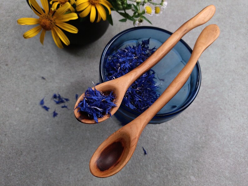 May include: Two light brown wooden spoons with smooth, curved handles. One spoon is filled with dried blue cornflower petals, while the other is empty, revealing a dark wood grain pattern in the bowl. The spoons are positioned over a blue glass bowl.