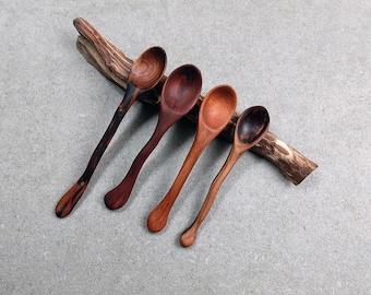 May include: Four handcrafted wooden spoons with distinct grain patterns and varying shades of brown, ranging from dark espresso to light amber, are arranged in a row resting against a piece of natural, textured driftwood.