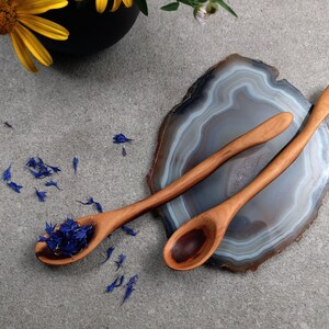 May include: Two light brown wooden spoons with smooth, elongated handles. One spoon contains a small pile of dried blue cornflower petals, while the other is empty. The spoons are positioned on a polished, translucent gray and white agate slice.