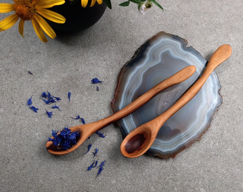 May include: Two light brown wooden spoons with smooth, elongated handles. One spoon contains a small pile of dried blue cornflower petals, while the other is empty. The spoons are positioned on a polished, translucent gray and white agate slice.