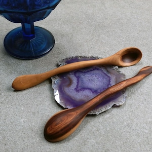 May include: Two wooden spoons with curved handles resting on a circular purple and white agate slice. One spoon is light brown with a smooth finish, while the other is a darker brown with visible wood grain patterns. A blue glass bowl is nearby.