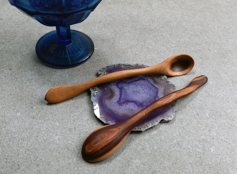 May include: Two wooden spoons with curved handles resting on a circular purple and white agate slice. One spoon is light brown with a smooth finish, while the other is a darker brown with visible wood grain patterns. A blue glass bowl is nearby.