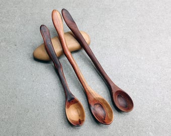 May include: Three long-handled wooden spoons with varying natural wood grain patterns and colors, ranging from light tan to deep dark brown, resting on a smooth, oval-shaped tan stone. Each spoon features a curved handle and a rounded bowl.