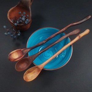 Op de afbeelding: Drie houten lepels met een lange steel en variërende natuurlijke houtnerven in tinten van lichtbruin tot donkerbruin, rustend in een fel turquoise keramische kom met een gladde, glanzende afwerking aan de binnenzijde.