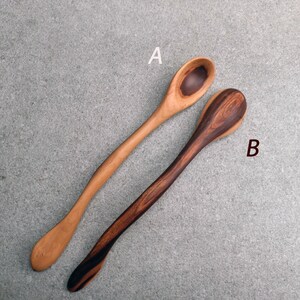 May include: Two wooden spoons with long, curved handles. Spoon A is light brown with a darker circular grain pattern in the bowl. Spoon B is dark brown with a prominent, swirling grain pattern throughout the handle and bowl. Both spoons are smooth.