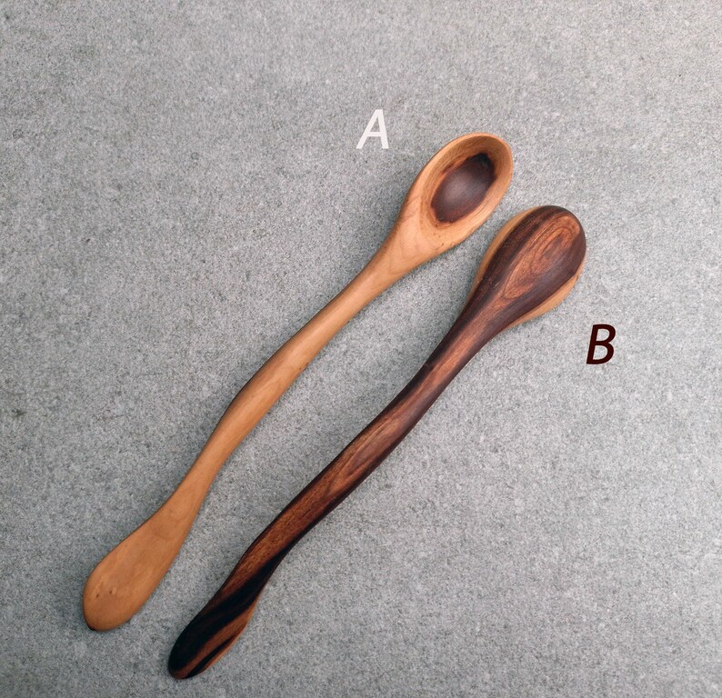 May include: Two wooden spoons with long, curved handles. Spoon A is light brown with a darker circular grain pattern in the bowl. Spoon B is dark brown with a prominent, swirling grain pattern throughout the handle and bowl. Both spoons are smooth.