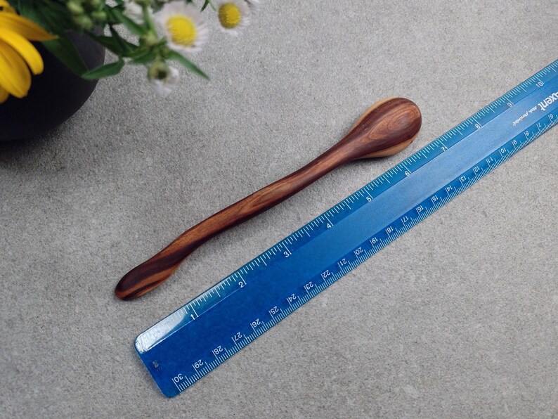 May include: A smooth, dark brown wooden spoon with a curved handle, positioned next to a blue plastic ruler that shows the spoon measures approximately 7.5 inches in length. The wood features a distinct grain pattern with lighter and darker streaks.