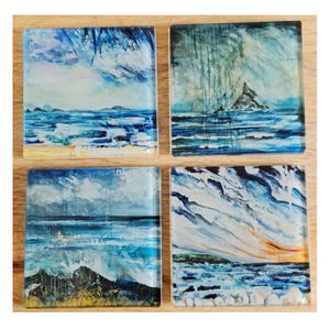 May include: Four square glass coasters with ocean landscape paintings. Each coaster features a unique abstract seascape in shades of blue, white, and yellow, depicting waves, sky, and land. The coasters are arranged on a wooden surface.