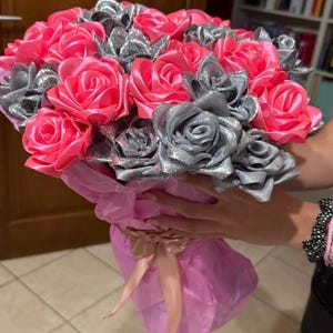 Può includere: Un bouquet di rose di stoffa fatte a mano, rosa e argento. Le rose sono disposte a bouquet e avvolte in carta velina rosa, legate con un nastro di raso. Il bouquet è tenuto da una persona.
