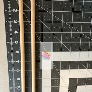 May include: Two wooden dowels, approximately 12 inches long, are shown against a black and white grid background. The dowels are natural wood color and are positioned vertically.