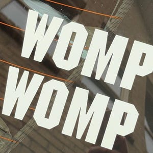 Può includere: Un adesivo in vinile bianco con la parola "WOMP" scritta due volte, una sopra l'altra. L'adesivo è su un finestrino di un'auto.