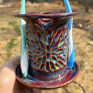 May include: A cylindrical ceramic toothbrush holder with a deep red and blue glaze featuring a textured, honeycomb-like pattern on the exterior. The holder has a wide base and two upright toothbrushes, one light green and one blue, inserted.
