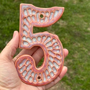 May include: A ceramic house number five in a peachy orange color with a white glaze. The number has a textured surface and two holes for hanging.