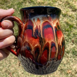 May include: A ceramic mug with a black rim and handle. The mug has a red, orange, and black glaze with a textured surface.