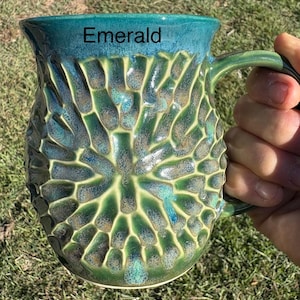 May include: A ceramic mug with a textured, honeycomb-like pattern in shades of emerald green and deep blue. The word Emerald is printed in black near the top rim. The mug features a curved handle and a glossy finish with varied color gradients.