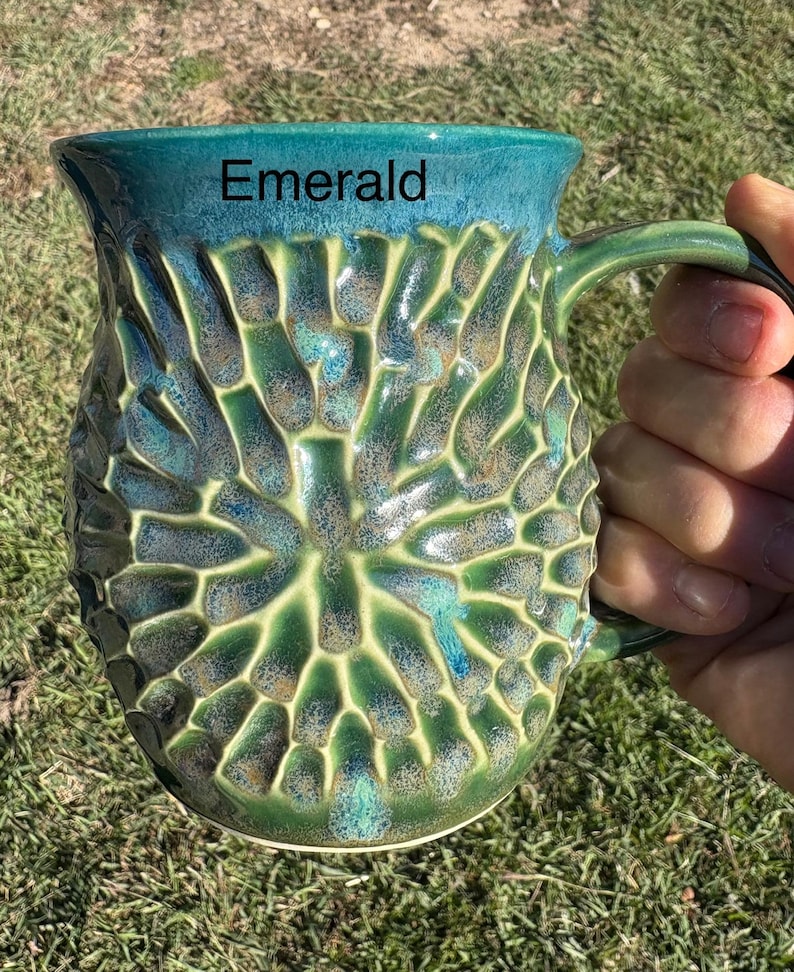 May include: A ceramic mug with a textured, honeycomb-like pattern in shades of emerald green and deep blue. The word Emerald is printed in black near the top rim. The mug features a curved handle and a glossy finish with varied color gradients.
