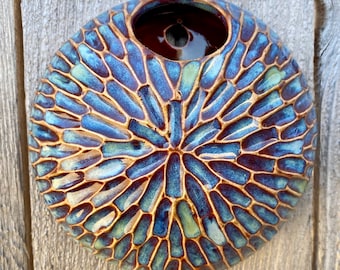 May include: A circular ceramic wall planter featuring a textured, radial pattern of raised, elongated segments. The surface is finished with a glossy glaze in shades of deep blue, light blue, and sage green, with a dark brown interior opening.