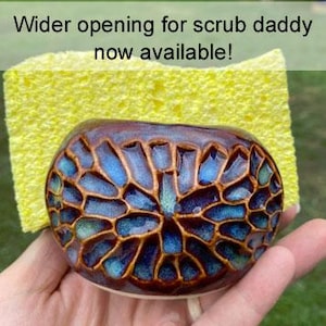 May include: A ceramic sponge holder with a brown and blue honeycomb-like textured pattern, holding a yellow rectangular sponge. The text on the image reads: Wider opening for scrub daddy now available!