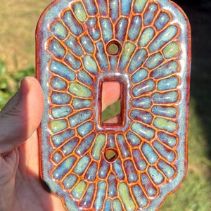 May include: A rectangular ceramic light switch plate with rounded, scalloped edges. The surface features a raised, pebble-like pattern in shades of blue and green, separated by deep, glossy reddish-brown grout lines and two screw holes.