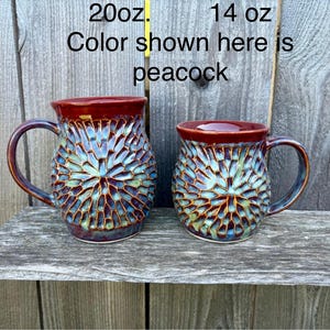 May include: Two ceramic mugs with a textured, radial pattern in blue, green, and brown, featuring a smooth dark red rim and handle. The text 20oz. and 14 oz. appears above the mugs, with the text Color shown here is peacock below.