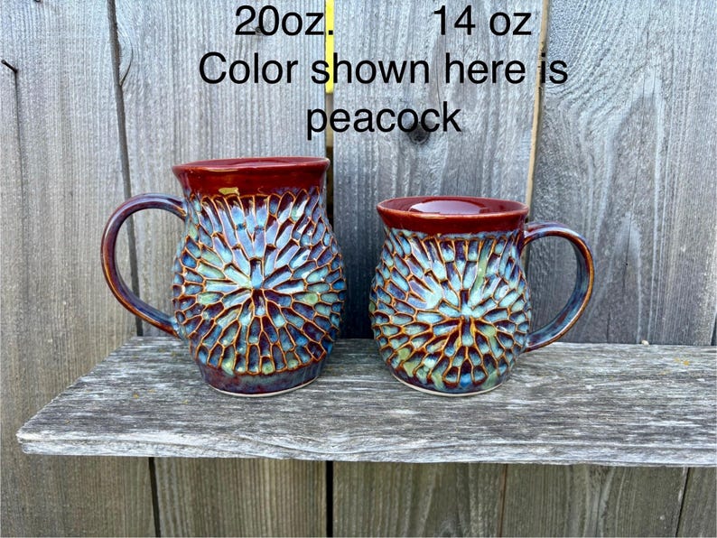 May include: Two ceramic mugs with a textured, radial pattern in blue, green, and brown, featuring a smooth dark red rim and handle. The text 20oz. and 14 oz. appears above the mugs, with the text Color shown here is peacock below.