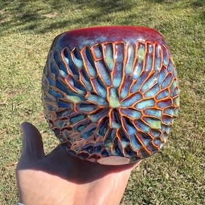 May include: A round ceramic bowl featuring a textured, honeycomb-like pattern with deep blue, teal, and light green glaze. The rim is finished in a dark reddish-brown tone, and the surface displays a glossy, multi-colored finish with orange-brown lines.