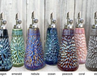 May include: A collection of seven ceramic oil bottles with stainless steel spouts. Each bottle features a unique color and textured design, including dragon, emerald, nebula, ocean, peacock, coral, and stone.