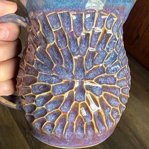 May include: A ceramic mug with a 14oz capacity labeled Nebula, featuring a textured, honeycomb-like pattern in shades of deep purple, blue, and brown. The glaze has a glossy finish with a smooth rim and a handle on the left side of the vessel.