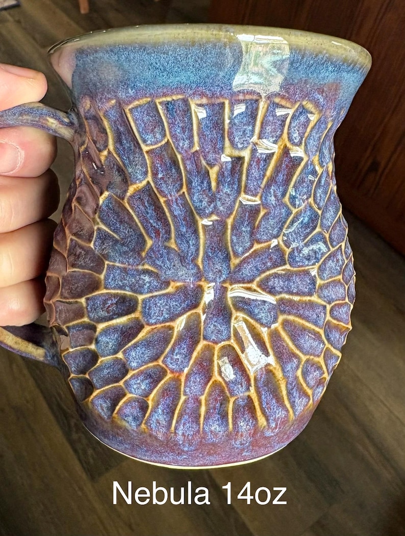 May include: A ceramic mug with a 14oz capacity labeled Nebula, featuring a textured, honeycomb-like pattern in shades of deep purple, blue, and brown. The glaze has a glossy finish with a smooth rim and a handle on the left side of the vessel.