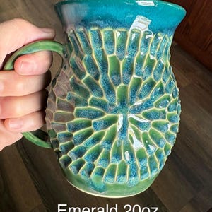 May include: A ceramic mug with a 20oz capacity, featuring a textured, honeycomb-like pattern on the exterior. The glaze transitions from a deep emerald green at the base to a vibrant teal at the rim. The mug includes a curved handle on the side.