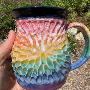May include: A ceramic mug featuring a textured, honeycomb-like carved surface with a vibrant rainbow gradient glaze. The colors transition from deep purple at the base to blue, green, yellow, orange, and pink, topped with a solid black rim and handle.
