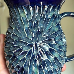 May include: A 20oz ceramic mug featuring a deep blue and teal glaze with a textured, radial carved pattern radiating from the center. The surface has a glossy finish with high-contrast highlights on the ridges and darker tones in the crevices.