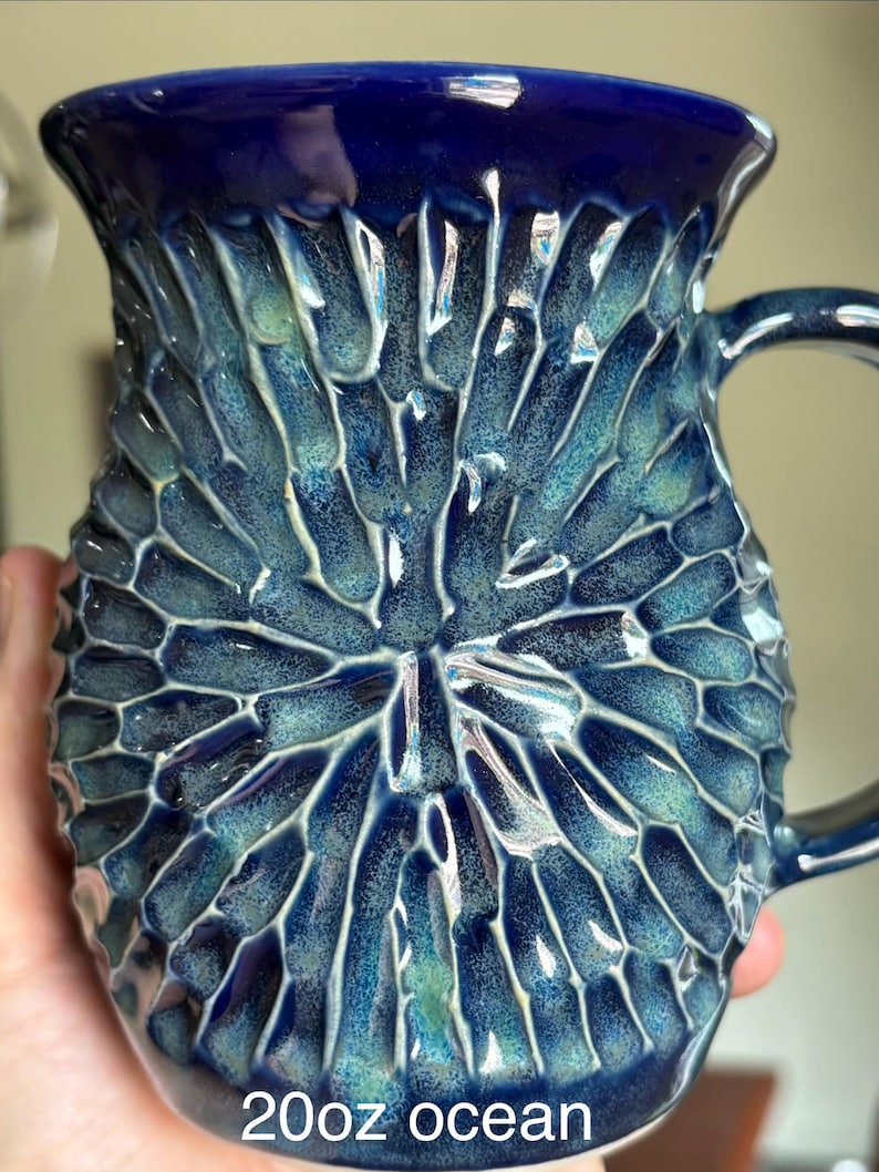 May include: A 20oz ceramic mug featuring a deep blue and teal glaze with a textured, radial carved pattern radiating from the center. The surface has a glossy finish with high-contrast highlights on the ridges and darker tones in the crevices.