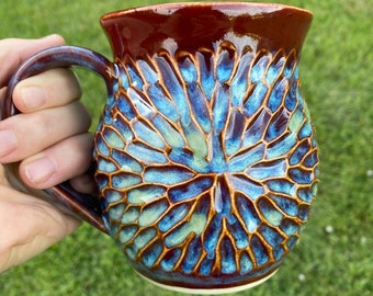 May include: A ceramic mug with a glossy brown rim and handle, featuring a textured, carved pattern on the body. The surface displays a vibrant blend of deep blue and light green glaze, creating a radial, petal-like design across the exterior.