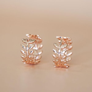 May include: Two rose gold hoop earrings with a leaf design. The earrings are displayed on a light brown surface. The text "Crush On Gold" is at the bottom of the image.