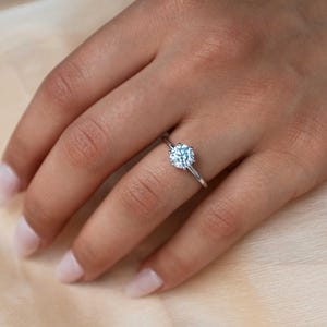 May include: A silver ring with a round, clear gemstone. The ring is on a finger, set against a soft, neutral-toned background. The gemstone is the focal point, reflecting light.