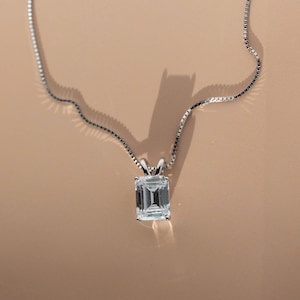 May include: A silver necklace with a rectangular, clear gemstone pendant. The pendant is set in a silver prong setting and hangs from a delicate silver chain. The background is a warm, neutral tone.