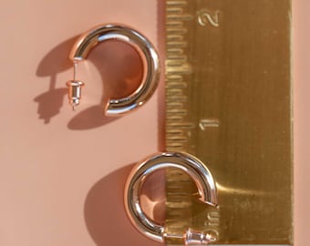 May include: Two gold-colored hoop earrings with a shiny finish are displayed on a pink surface. A ruler is placed next to the earrings. The earrings are approximately 0.75 inches in diameter, showcasing a classic jewelry design.