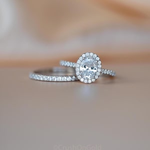 May include: A silver engagement ring set with a large oval-shaped center stone surrounded by a halo of smaller stones. The band is also set with smaller stones.