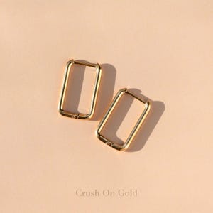 May include: Two gold-colored rectangular hoop earrings. The earrings are displayed on a light peach-colored surface. The text "Crush On Gold" is at the bottom of the image.