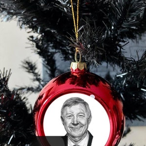 Puede incluir: Una brillante bola roja de Navidad con un retrato de un hombre sonriente. La bola cuelga de un hilo dorado contra ramas negras de árbol de Navidad. El adorno tiene el texto "Gemma Firth FINE ART".