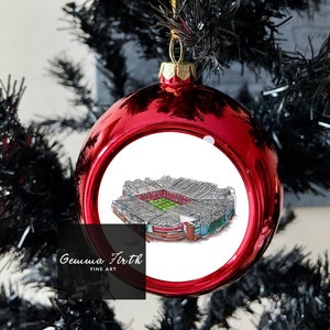 Puede incluir: Una bola de Navidad roja brillante con una tapa y un cordón dorados, colgada de un árbol de Navidad negro. La bola presenta una ilustración detallada de un estadio. El texto "Gemma Firth FINE ART" es visible.