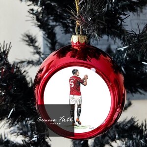 Puede incluir: Una bola de Navidad roja brillante con una tapa y cuerda doradas, colgando de un árbol de Navidad negro. La bola presenta una ilustración de un futbolista con una camiseta roja con el número 5. El texto "Gemma Firth FINE ART" es visible.