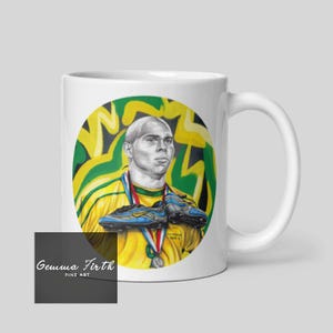 Puede incluir: Taza de cer&aacute;mica blanca con una ilustraci&oacute;n colorida de un jugador de f&uacute;tbol sosteniendo dos botas de f&uacute;tbol. El jugador lleva una camiseta verde y amarilla con una medalla de oro alrededor del cuello. El fondo es un dise&ntilde;o de graffiti verde y amarillo. La taza tiene el texto "Gemma Firth Fine Art" en la parte inferior.
