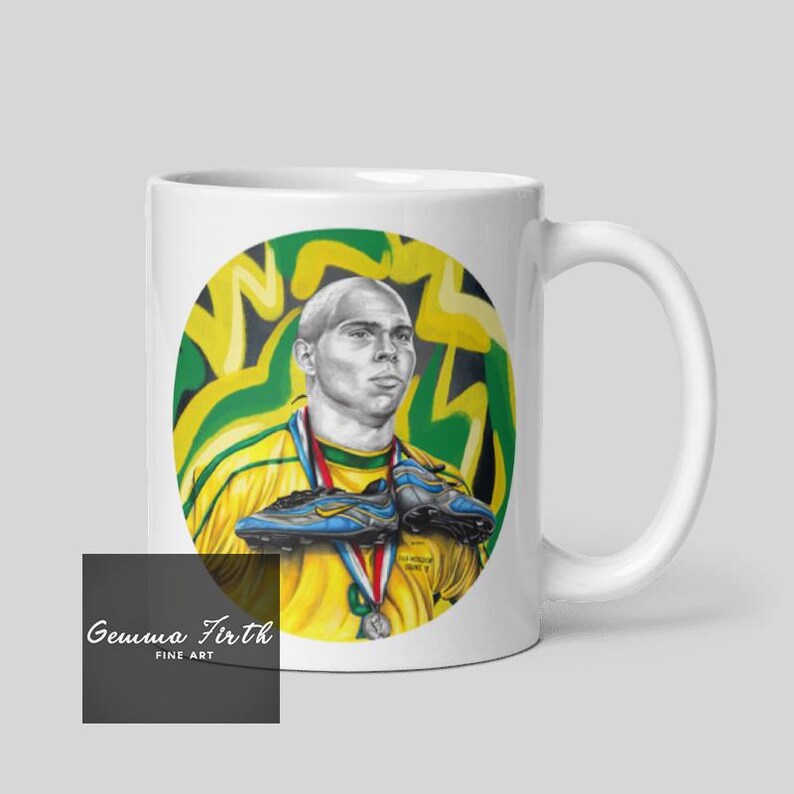 Puede incluir: Taza de cer&aacute;mica blanca con una ilustraci&oacute;n colorida de un jugador de f&uacute;tbol sosteniendo dos botas de f&uacute;tbol. El jugador lleva una camiseta verde y amarilla con una medalla de oro alrededor del cuello. El fondo es un dise&ntilde;o de graffiti verde y amarillo. La taza tiene el texto "Gemma Firth Fine Art" en la parte inferior.