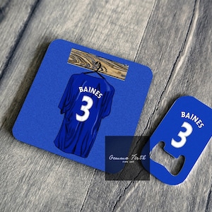 Könnte beinhalten: Blau-weißes Trikot des Everton Football Clubs mit dem Namen "BAINES" und der Nummer "3" auf der Rückseite, das an einem Holzaufhänger hängt. Das Trikot befindet sich auf einem blauen Untersetzer mit einem Flaschenöffner.