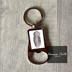 May include: A silver bottle opener keyring with a white rectangular image of a stadium. The image is surrounded by a black border. The keyring has a silver ring and the text "Gemma Firth Fine Art" is printed on the bottom.