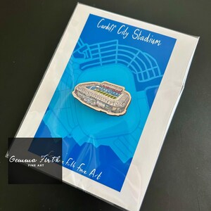 Peut inclure: Épinglette en bois représentant le stade de Cardiff City, avec des détails architecturaux, sur un fond bleu avec un motif de carte. Le texte "Cardiff City Stadium" est au-dessus de l'épinglette. L'œuvre est présentée sur une carte blanche.